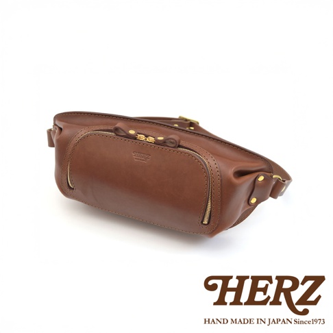 HERZ 品牌斜背包，採用棕色皮革製作，主體設計簡潔，帶有雙拉鍊前袋和金屬鉚釘細節，展現手工製作質感。