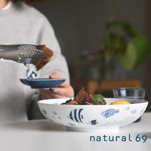 一個裝有食物的白色陶瓷大圓深碗，碗身飾有藍色魚和珊瑚圖案。一位身穿灰色毛衣的人正用夾子將肉放入碗中。