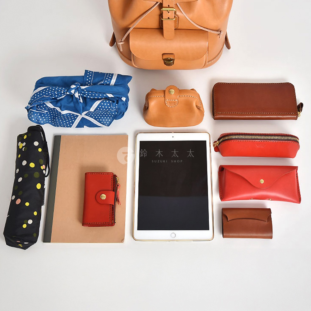 多個皮革配件，包括一個淺棕色皮革後背包、一個棕色皮革長夾、一個紅色皮革筆袋、一個紅色皮革零錢包、一個紅色皮革眼鏡盒、一個棕色皮革卡夾，以及一個帶有彩色圓點的黑色摺疊傘、一個藍色印花頭巾和一本筆記本。