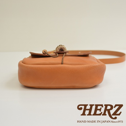 棕色皮革製成的職人手作肩背包，特色為鹿角扣和細皮革繩束口設計，包身圓潤飽滿，附有可調整的皮革長背帶。