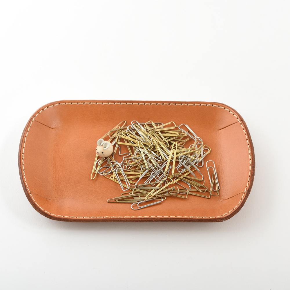 職人手作皮革小物收納盤，淺棕色皮革，邊緣縫線，盤中裝有大量銀色與金色迴紋針，左側有一個可愛的木製小老鼠裝飾。