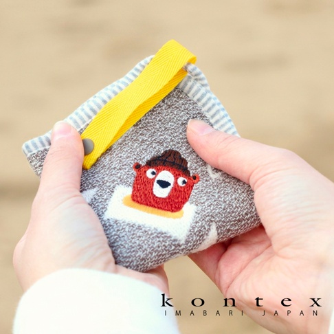 一雙手正拿著一個方形的 KONTEX 今治純棉方巾，布料呈現灰白色織紋，中間繡有一個戴著咖啡色毛帽的紅色小熊圖案，頂部附有一個明黃色的織帶掛環與灰色暗釦，背景為模糊的淺色調。