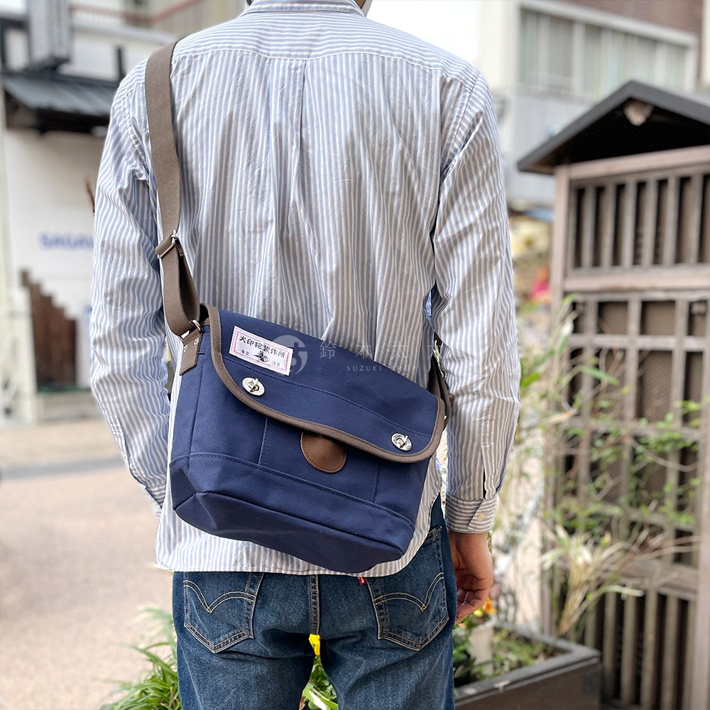 深藍色純綿帆布雙扣郵差包，配有棕色皮革飾邊和同色系肩帶，包蓋上有日文標籤。