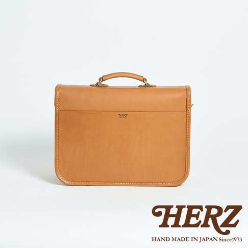 焦糖色皮革手提包，方形設計，頂部有提把和金屬扣環。包身正面有一個壓印的品牌標誌，縫線精緻，質感厚實。