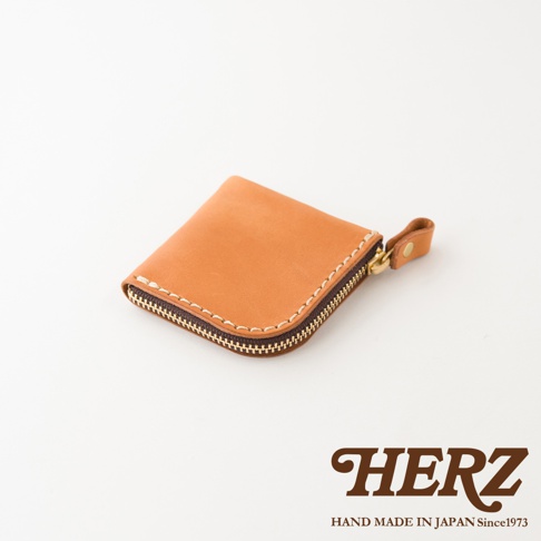 一個日本HERZ手工製作的皮革零錢包，呈現焦糖色調，採用紮實的皮革材質與對比色縫線，側面配有金屬拉鍊與皮革拉環，整體外型簡約精緻，右下角印有HERZ品牌商標。