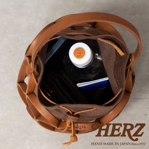 日本HERZ職人手作皮革Organ編織水桶包，內部裝有水瓶、皮夾和藍色筆記本，包款採皮革編織設計，以俯視角度拍攝。