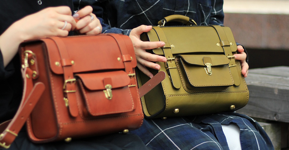 兩款學院風迷你後背包，一款為棕褐色皮革，另一款為橄欖綠皮革，皆有雙翻蓋設計、金色金屬扣環與銅色卯釘裝飾，搭配可拆卸肩帶。