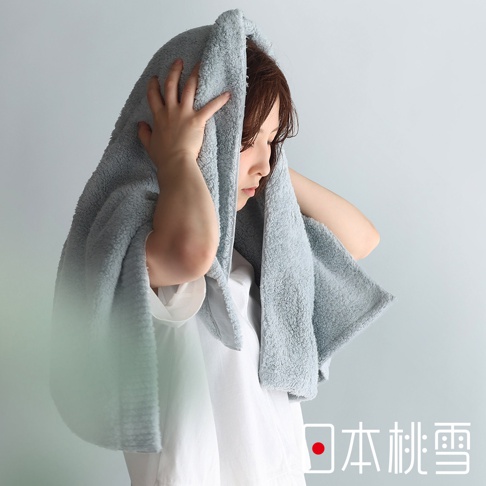 一名女子用一條淺藍色的蓬鬆毛巾包裹住頭部，毛巾的材質看起來非常柔軟，她雙手正將毛巾整理好，臉部表情專注，背景是淺灰色的牆壁。圖片右下方有「日本桃雪」的日文標誌。