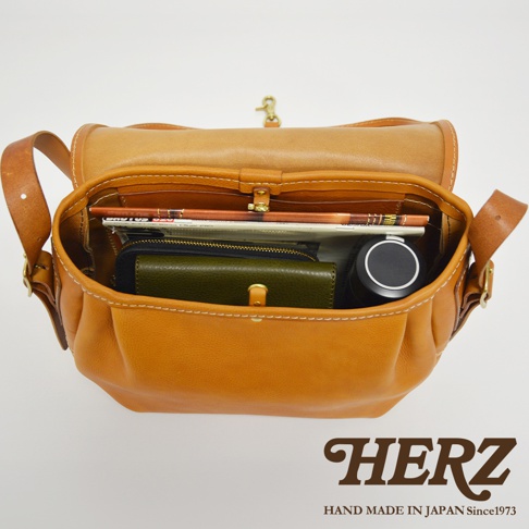 職人手作皮革復古翻蓋郵差包，可見內部裝有雜誌、綠色皮夾、黑色圓筒狀物品，包身為淺棕色皮革，縫線明顯。