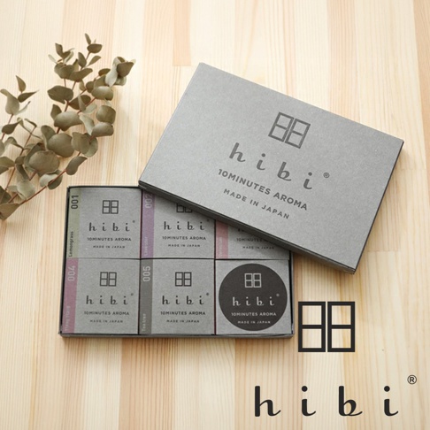 木紋桌面上擺放一盒打開的 hibi 香氛火柴禮盒，內部整齊排列五個標註數字的小型香氛盒及專用燃燒墊，上方覆蓋著印有 hibi 品牌標誌與文字的灰色包裝盒蓋，左側搭配一株綠色尤加利葉裝飾。