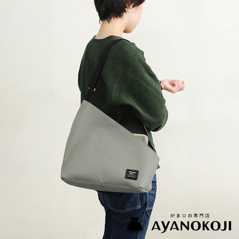 一名女子身穿綠色毛衣，斜揹著一個灰色方形口金單肩包，包上有品牌的黑色標籤，背景為純白色。