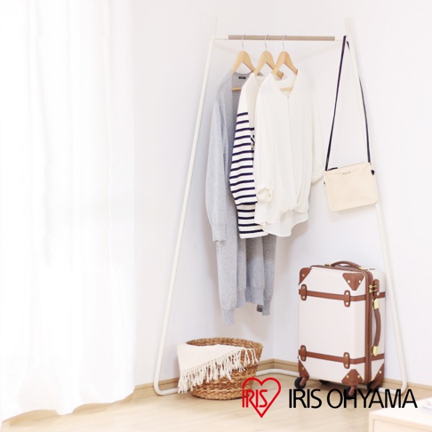 白色角落造型吊掛衣桿，上掛有灰色長袖外套、條紋長袖上衣和白色長袖上衣，旁邊掛著米白色斜背包，下方有竹編籃和復古風格行李箱。