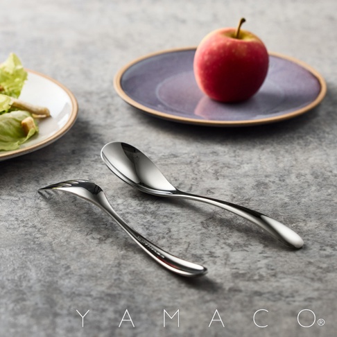 兩個不鏽鋼餐具，一個湯匙和一個叉子，擺放在灰色石板紋理的背景上。湯匙在後，叉子在前。背景後方有一個淺盤，盤上有一個紅蘋果。盤子左側還有一個小盤子，盤中有部分生菜沙拉。圖片下方印有YAMACO字樣。