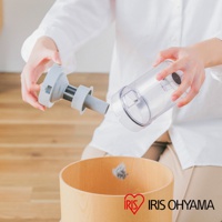 一位女性手持吸塵器集塵筒和濾網組件，準備將灰塵倒入木製垃圾桶。集塵筒為透明塑膠材質，濾網組件為灰色塑膠。