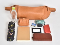職人手作皮革大容量斜背包，淺棕色皮革材質，搭配金色金屬扣環。包內物品包含：一瓶礦泉水、黑色圓點圖案摺疊傘、淺棕色筆記本、紅色皮革卡夾、黑色長夾、黑色無線耳機盒、淺綠色印花收納包及一張票券。