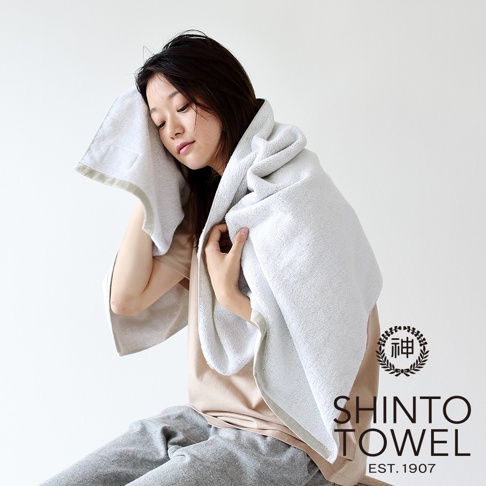 一位亞洲女性使用一條蓬鬆柔軟的淺灰色有機棉浴巾擦拭臉部，浴巾搭在肩上。