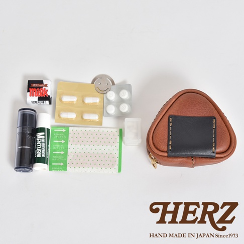 職人手作皮革小物收納包，為三角飯糰造型，主體為棕色皮革，正面縫有黑色皮革飾片，搭配金色拉鍊。收納包旁擺放有藥品、藥膏、貼布及一小盒藥丸。