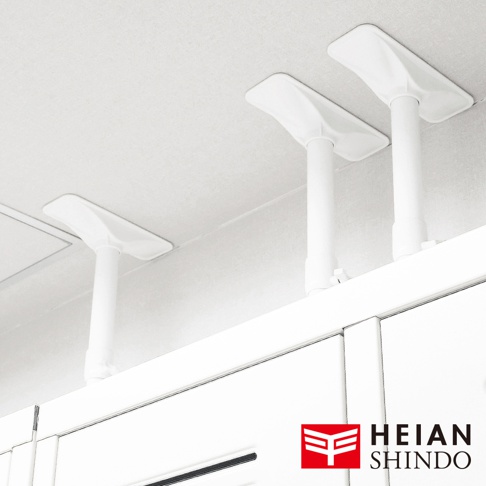 白色伸縮防傾桿，頂部有梯形固定墊，安裝於天花板與白色櫥櫃之間，展示了其支撐與固定功能。