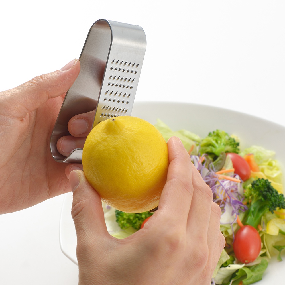 一隻手拿著日製立式磨泥器，另一隻手握住一顆黃色檸檬，準備磨取檸檬皮屑，下方是裝有生菜、花椰菜、小番茄等食材的沙拉。