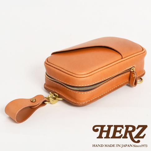 日本HERZ品牌手工皮革斜背包，棕色皮革，附有金色拉鍊和吊飾，文字標示HERZ HAND MADE IN JAPAN Since1973