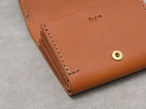 職人手作皮革Organ雙折迷你短夾，呈現溫潤的棕褐色調，皮革表面壓印「Organ」字樣，並配有金色按扣。