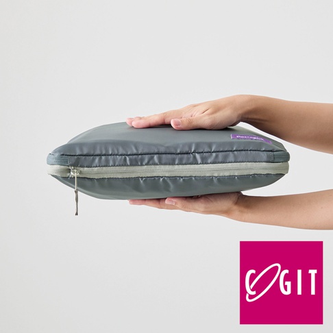 一雙手正拿著一個灰色的折疊壓縮旅行袋，袋子為扁平狀，拉鍊處有淺灰色滾邊，袋子上印有紫色標籤。