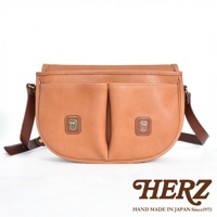 職人手作皮革傳統釣魚包，呈現自然的淺棕色調。包身為圓弧形設計，正面有兩個對稱的貼袋，每個貼袋上各有一個帶有金屬扣的方形皮革飾片。包包配有深棕色的皮革肩帶。