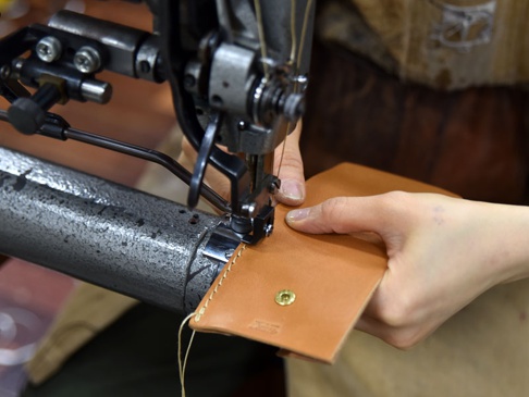 職人手作皮革雙折收納零錢包，淺棕色皮革，由工業縫紉機縫製中，可見金色按扣。