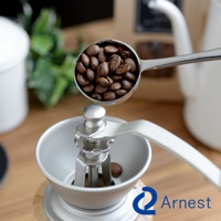 銀色金屬咖啡量匙夾，裝滿烘焙過的咖啡豆，正上方對準一台手搖咖啡磨豆機的漏斗口。