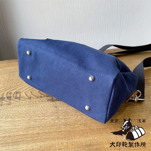 深藍色帆布側背包底部，設有四個銀色金屬腳釘，背包帶為黑色，圖片右下角有日文品牌標誌