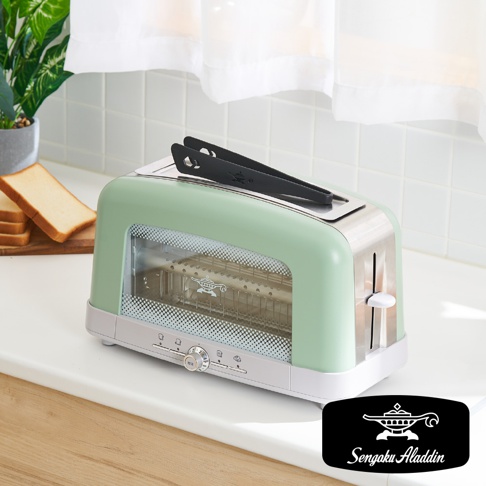 淺綠色搭配銀色不鏽鋼邊框的烤麵包機，配有黑色夾子，機身側面有透明玻璃視窗，底部有旋鈕和按鈕。