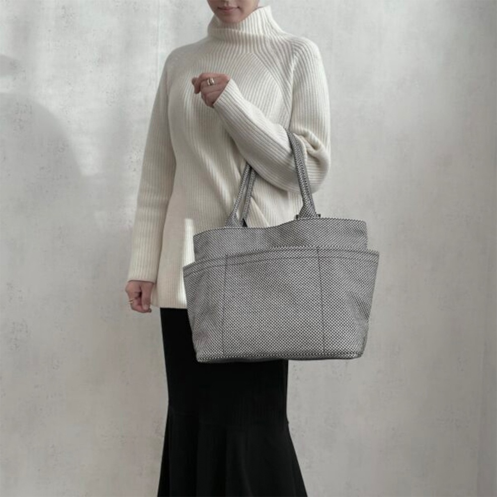 一位女士手提一個灰色刺子織紋理托特包，包身有明顯的格紋細節與多個外袋。