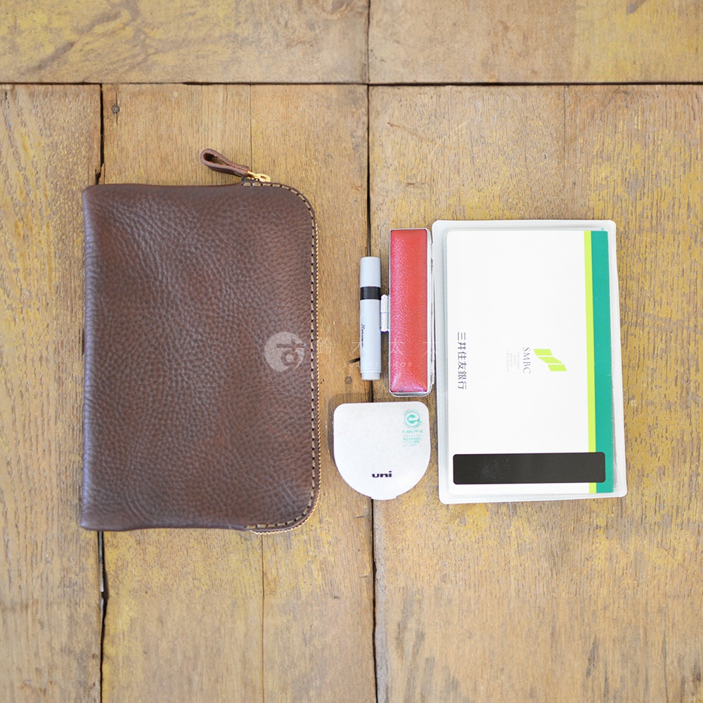 職人手作皮革Organ雙拉鍊收納袋，深棕色紋理皮革，配有金色拉鍊。旁邊放置著一個白色小型印章、一個紅色印章盒，以及一本白色封面、綠色邊條的存摺。