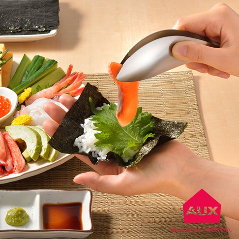 手持不鏽鋼調理夾，夾取鮭魚片，準備製作壽司，旁邊擺有紫蘇葉、白飯、海苔、酪梨、小黃瓜、蝦子、魚卵及醬油芥末。