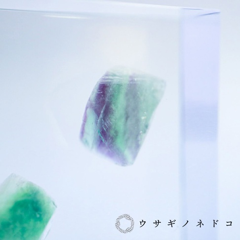 一塊1.5公分厚切型片的螢石標本，呈現紫色與綠色的條紋，置於透明展示盒中，背景為淺藍色調。