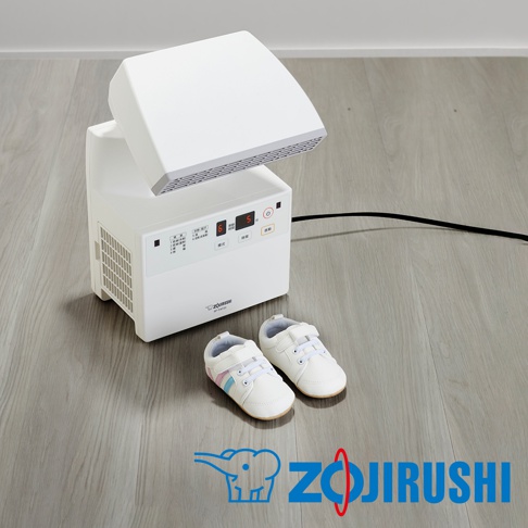 白色多功能智慧烘乾機，設有 LED 顯示螢幕和控制按鈕，上方頂蓋可向上傾斜，旁邊地面放著一雙白色嬰兒鞋，鞋上有粉色和藍色條紋裝飾。