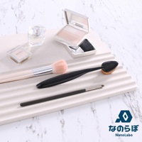 淺色珪藻土置物墊上擺放著三支化妝刷（一支圓形粉刷，一支黑色扁平刷，一支細長眼影刷）、一瓶方形透明香水、一個方形透明粉餅盒，以及一個白色粉餅盒，盒內有粉餅和粉撲。