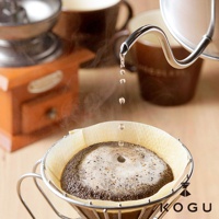 日製18-8不鏽鋼極細口手沖壺，壺嘴正將水滴注入濾杯中的咖啡粉。背景有木質咖啡研磨機與咖啡杯。