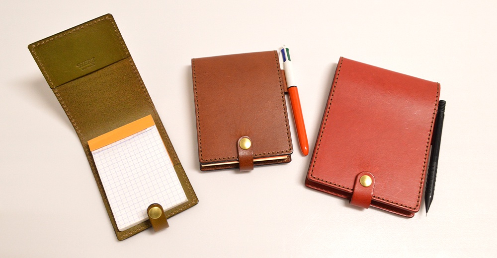 三款職人手作皮革便利貼收納套，分別為綠色、棕色和紅色，均採用皮革材質，配有金屬按扣。其中綠色款打開展示了內部的便利貼和筆槽，棕色和紅色款則有筆插入其中。