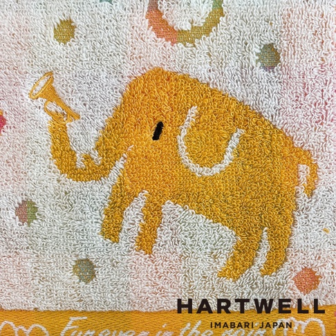 這是一條 HARTWELL 今治毛巾，純白色的毛巾表面印有淡彩圓點圖案，中央刺繡著一隻亮黃色的可愛小象，小象的鼻子向上捲起，手持一支黃色小號，整體風格簡約溫馨，右下角印有 HARTWELL IMABARI JAPAN 字樣。