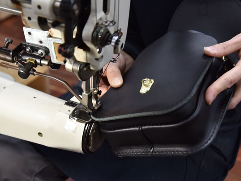 職人手作黑色皮革翻蓋迷你單肩包，包身上有金色旋鎖扣，正在由工業縫紉機縫製中。