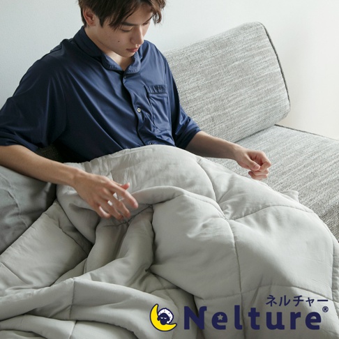 淺灰色 Nemulia 五層散熱涼感單人被，採用柔軟材質，表面有菱格紋縫製，覆蓋在一名身穿深藍色睡衣的男子身上，男子坐在灰色沙發上。