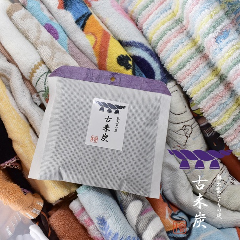 一個白色紙袋包裝的古來炭迷你調濕木炭，袋面印有日文標籤和圖案。背景是多條捲起的彩色毛巾。