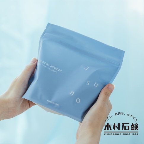 雙手捧著一袋淺藍色、霧面質感的 ASUNO 無香料溫和中性洗衣粉包裝。包裝上有白色字體標示「LAUNDRY POWDER」及日文說明，以及「a s u n o」的字母設計。