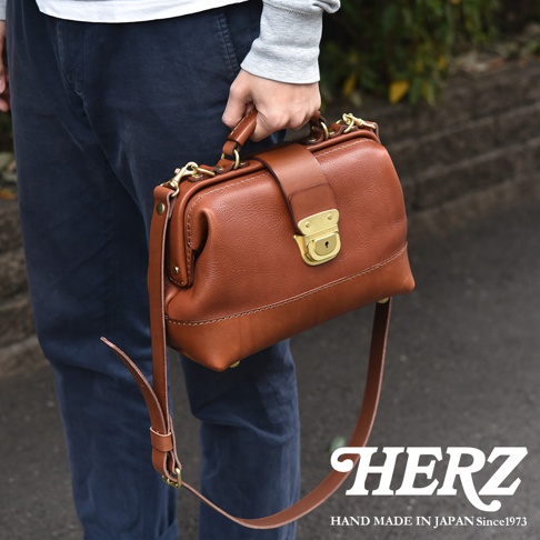 一位男子手持日本HERZ職人手作皮革兩用迷你口金包，包款呈現復古棕色皮革質感，搭配黃銅色金屬扣環與可拆卸式肩帶，展現細膩精緻的日系職人工藝。