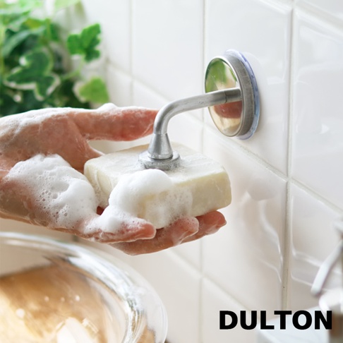 銀色金屬材質的磁吸式皂架，末端有一個尖頭，插入白色肥皂中，肥皂上佈滿泡沫，一隻手拿著肥皂，皂架吸附在白色磁磚牆上。