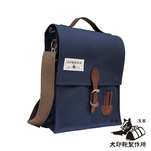 深藍色純棉帆布製成的直式側背包，配有棕色皮革提把、可調式斜背包帶，以及一個帶有金屬扣環的皮革翻蓋。