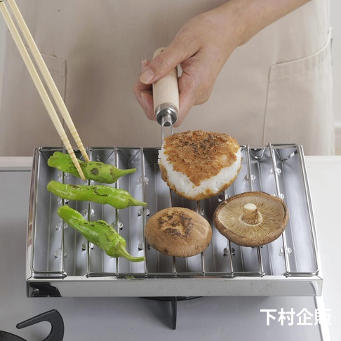 瓦斯爐專用炭烤風萬用燒烤盤，不鏽鋼材質，烤架上擺放著綠色的辣椒、香菇和烤飯糰，旁邊有雙筷子夾著辣椒。