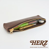 棕色皮革製的職人手作收納筆袋，拉鍊半開，內含一支綠色筆、一支黃銅筆和一塊橡皮擦。