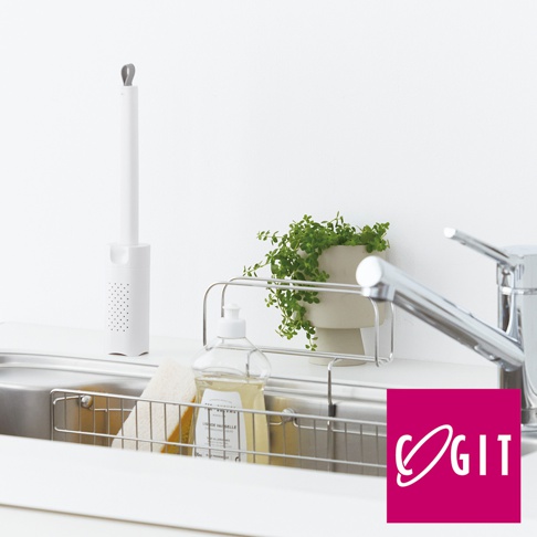 白色推拉式海綿刷，配有灰色環狀提把，下方為圓筒狀機身，帶有網狀透氣孔。旁邊是廚房水槽，內有不鏽鋼瀝水架，架上擺放著一瓶透明洗碗精和一個黃色海綿。背景是簡潔的白色牆面，角落擺放著一盆綠色植物，旁邊是銀色廚房水龍頭。